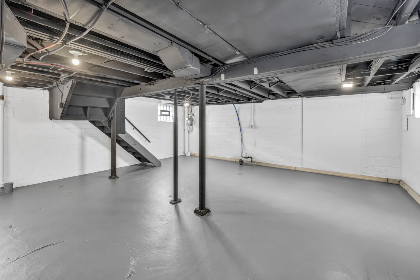 An unfinished basement with painted white block walls, steel support posts and a smooth grey concrete floor running to the wall joint.