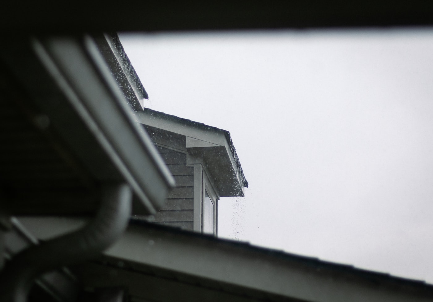 Rain running off a shingled roof edge and past a downspout on an overcast day, seen from beneath the eave.