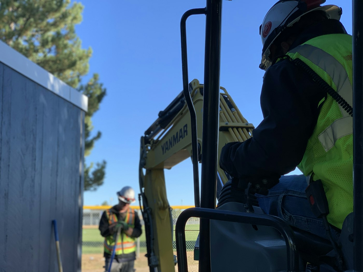 An operator sitting in the cab of a compact excavator with a second worker standing at the edge of the cut holding a shovel, on a clear morning.