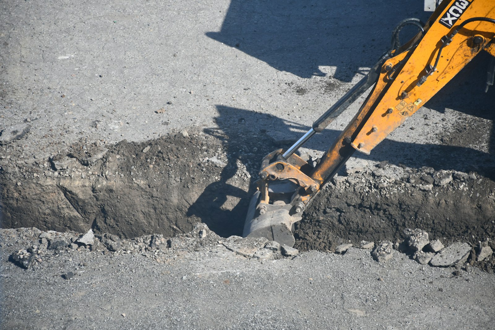 An excavator bucket set down into a narrow open cut, broken material stacked along both sides of the trench.